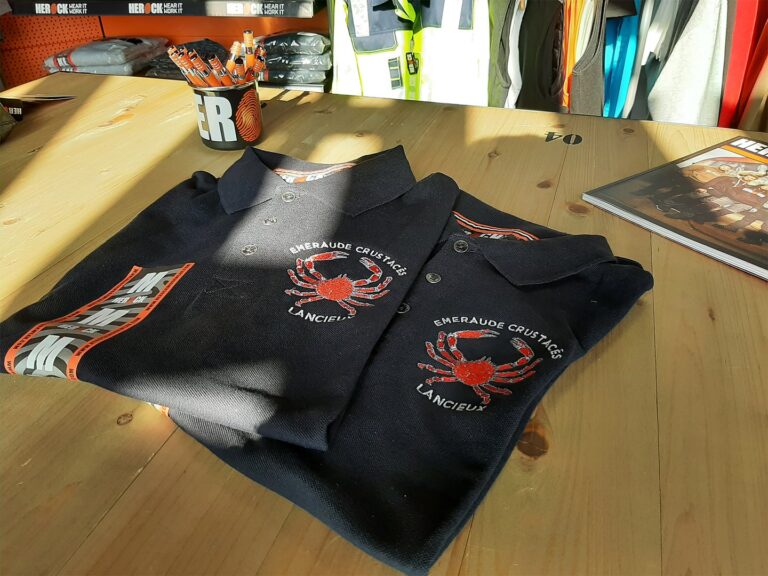 Polos noirs brodés avec logo crabe rouge Emeraude Crustacés Lancieux sur une table en bois.